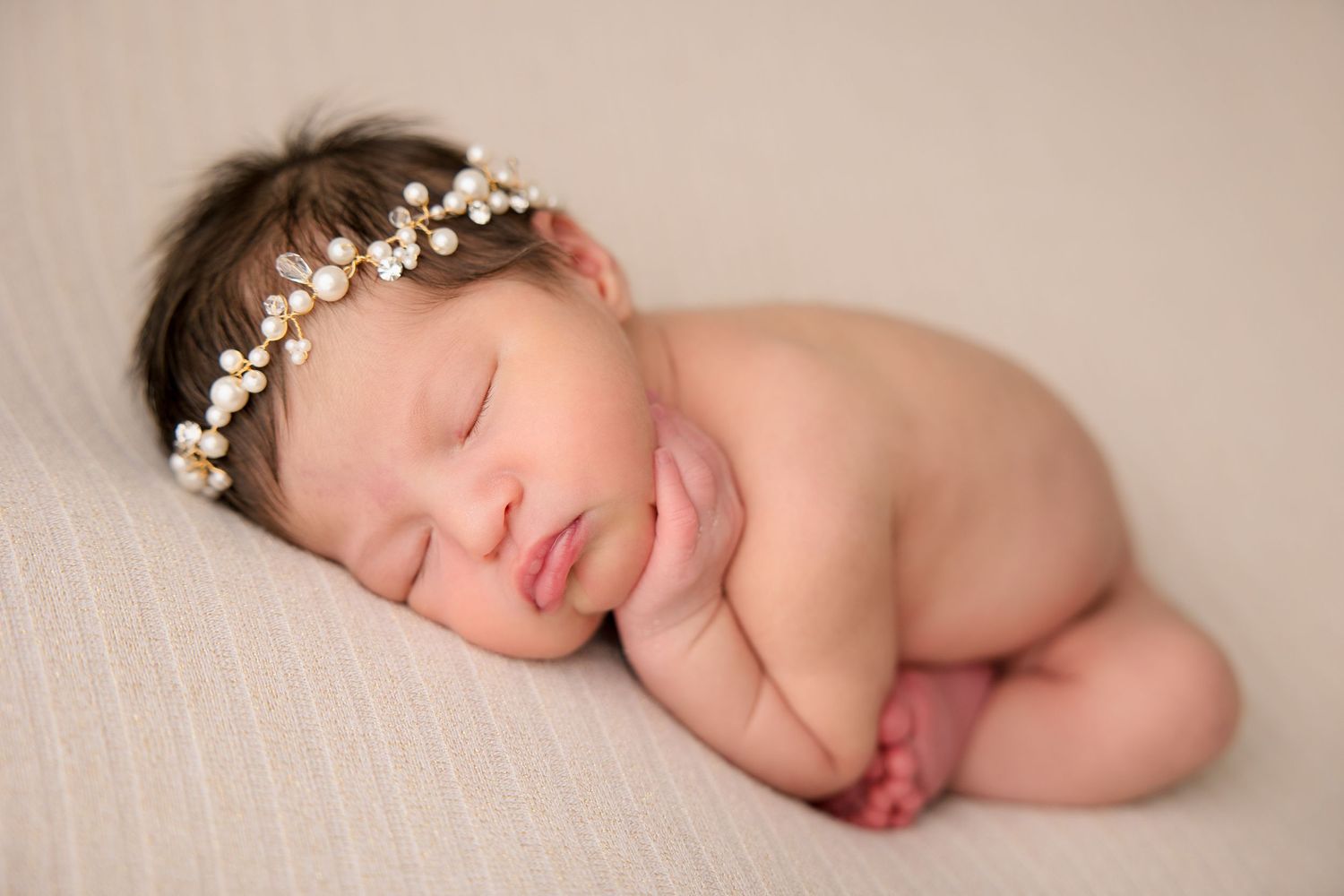 Sleeping newborn wearing delicate floral crown positioned on cream colored backdrop.