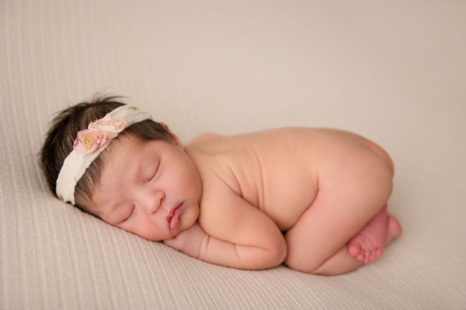 Sleeping newborn baby wearing floral headband on textured cream backdrop.