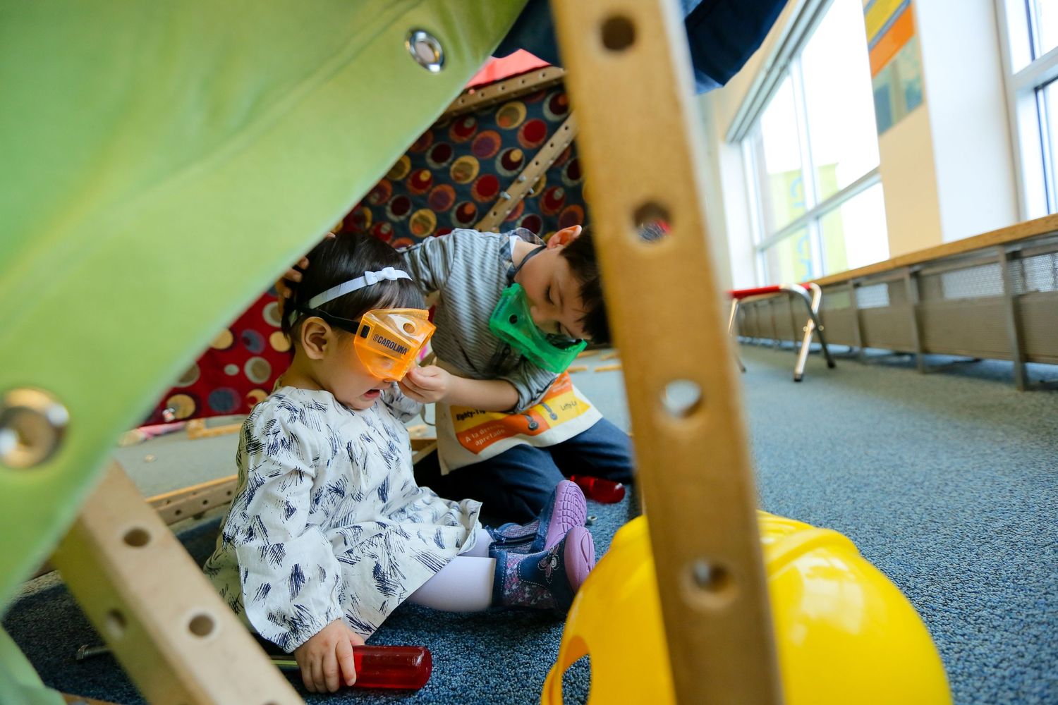 Kids playing with wooden construction toys on a carpeted floor in a bright room.