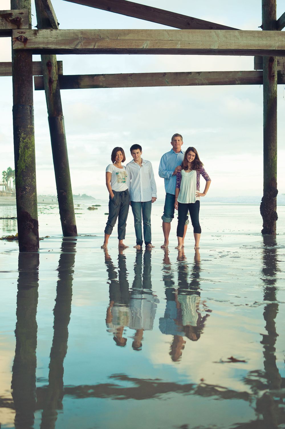 Family of four standing barefoot under a pier reflected in wet sand at low tide.
