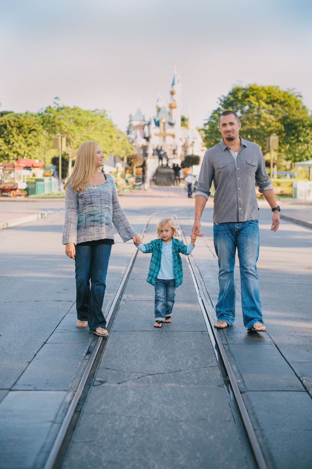 Family of three holding hands walking toward Sleeping Beauty Castle at Disneyland on a sunny day.