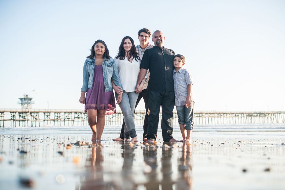 Family of five stands together on wet sand beach with pier in background.