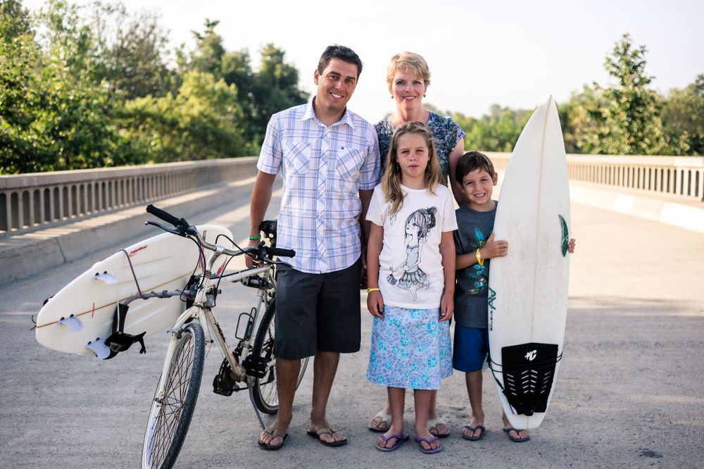 Family of four poses with bicycle and surfboard on sunny bridge outdoors.