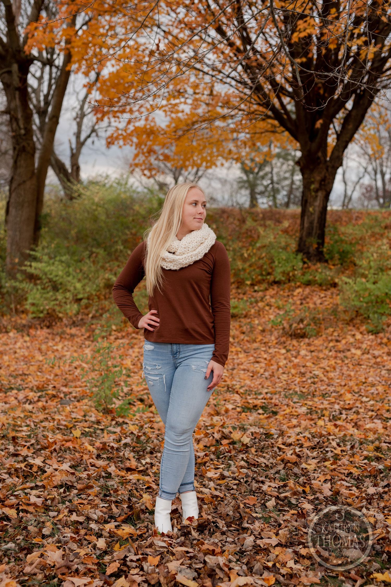 Person in brown sweater and white scarf posing against vibrant autumn trees.