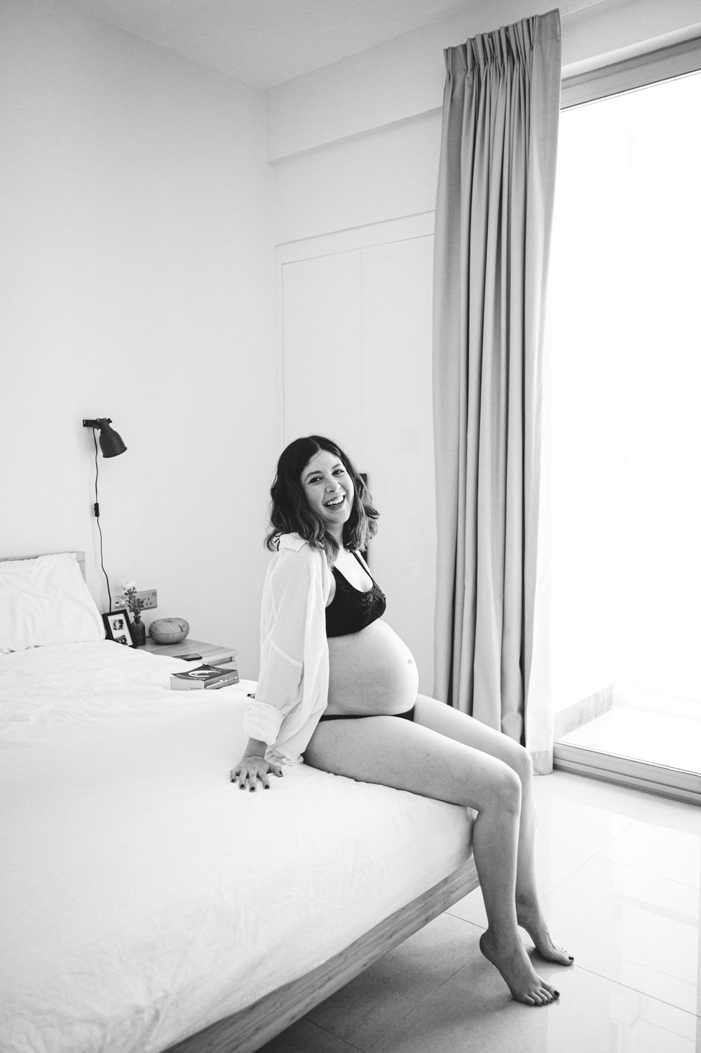 Black and white maternity photo shows relaxed pose on bed by window.