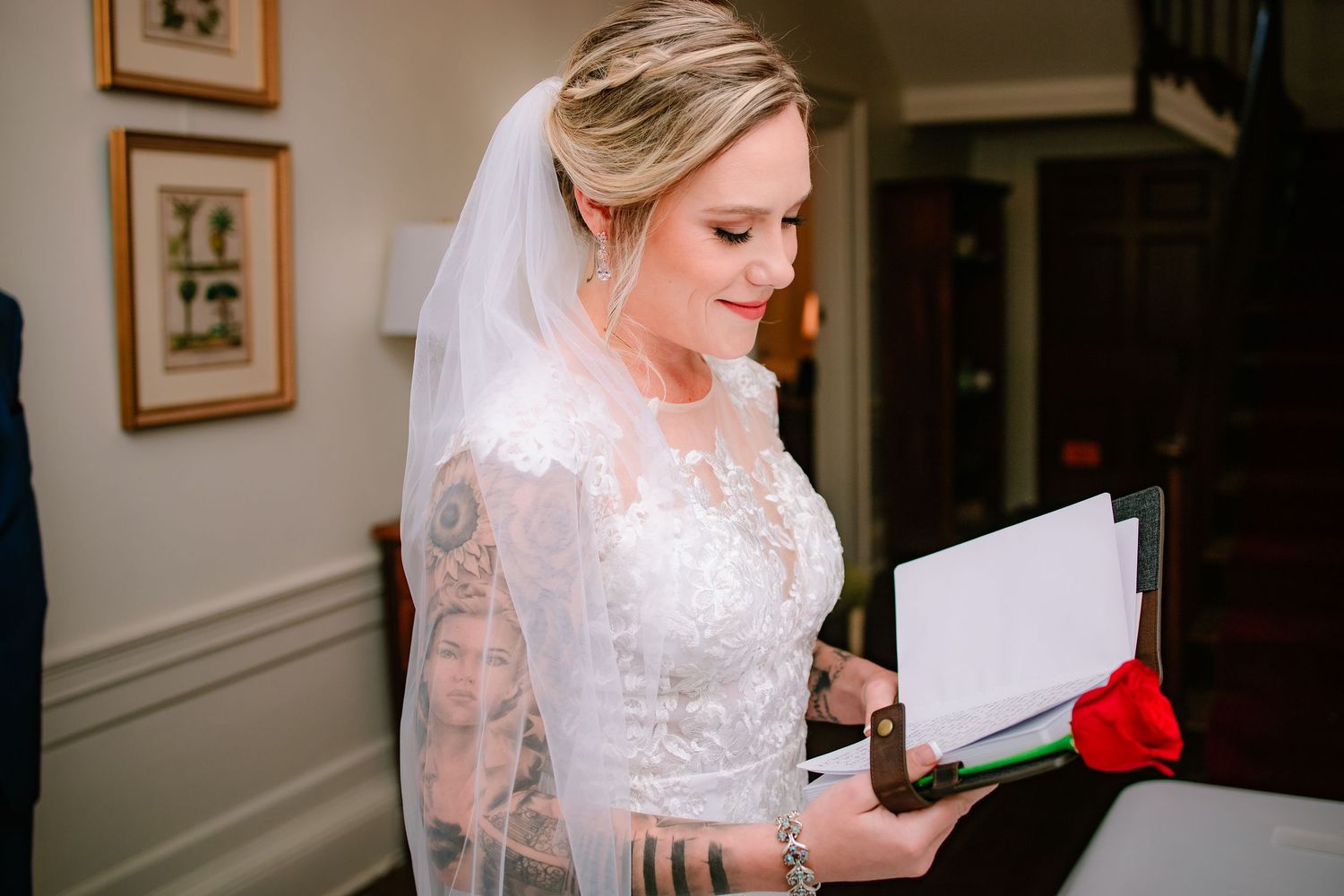 A tattooed bride in white lace long-sleeve gown reads a note while holding a red rose indoors before her wedding.