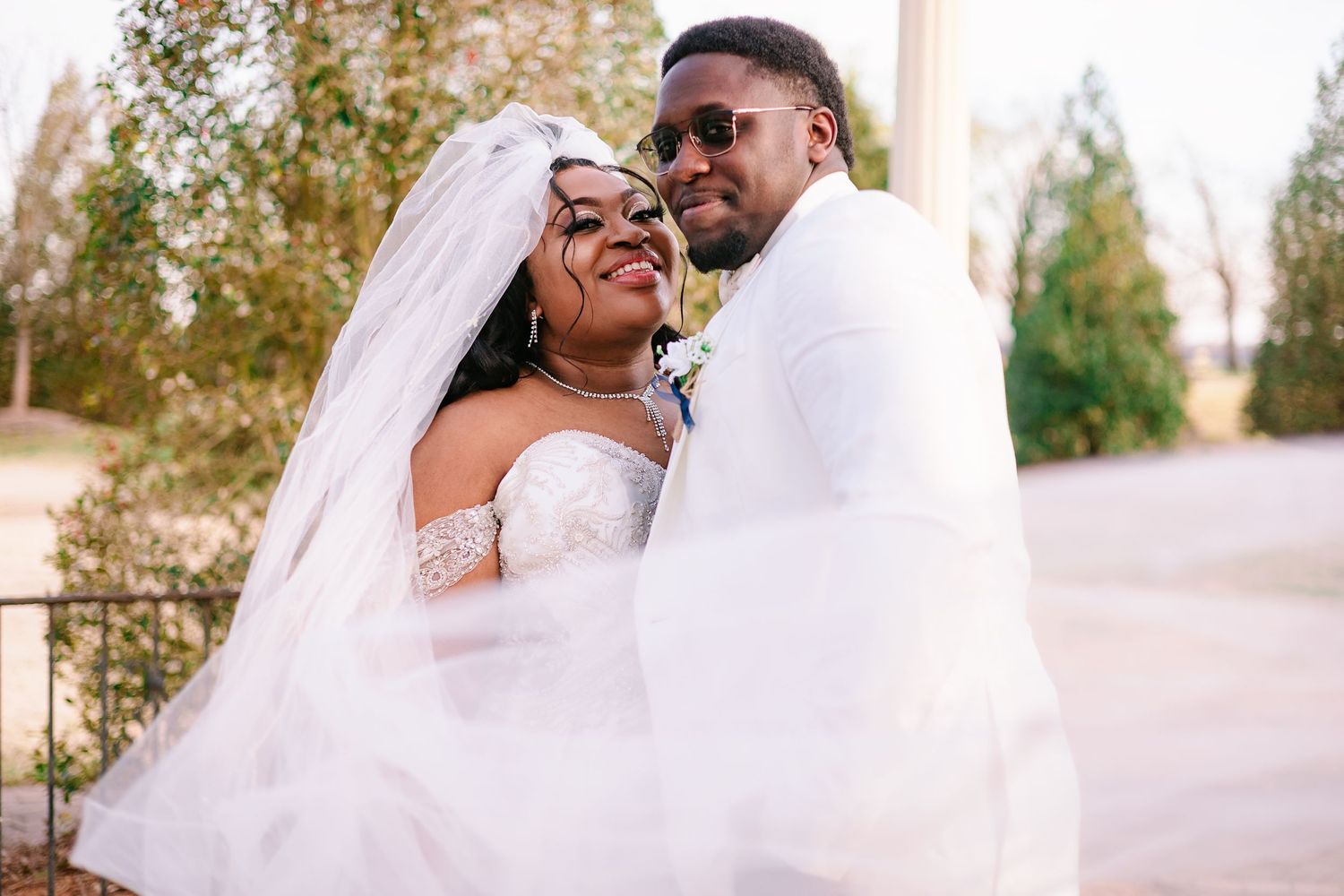 A happy Black newlywed couple posing together, bride in white lace gown with veil, groom in white suit wearing sunglasses.