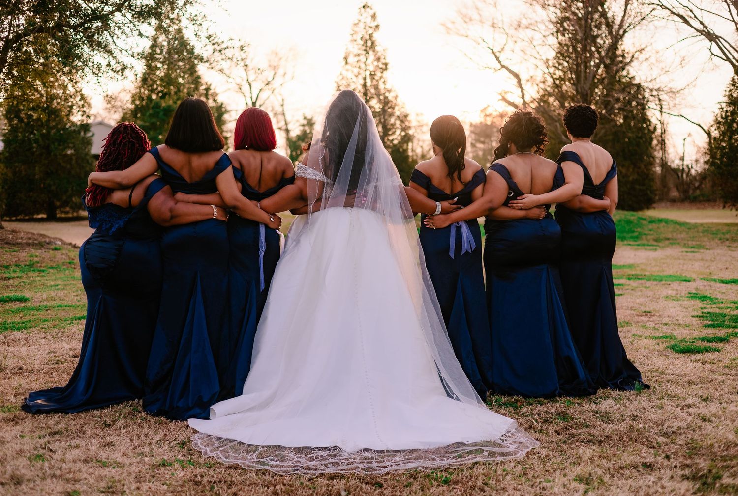 A bride in white wedding gown with veil stands with bridesmaids in navy blue dresses, all facing away at sunset.