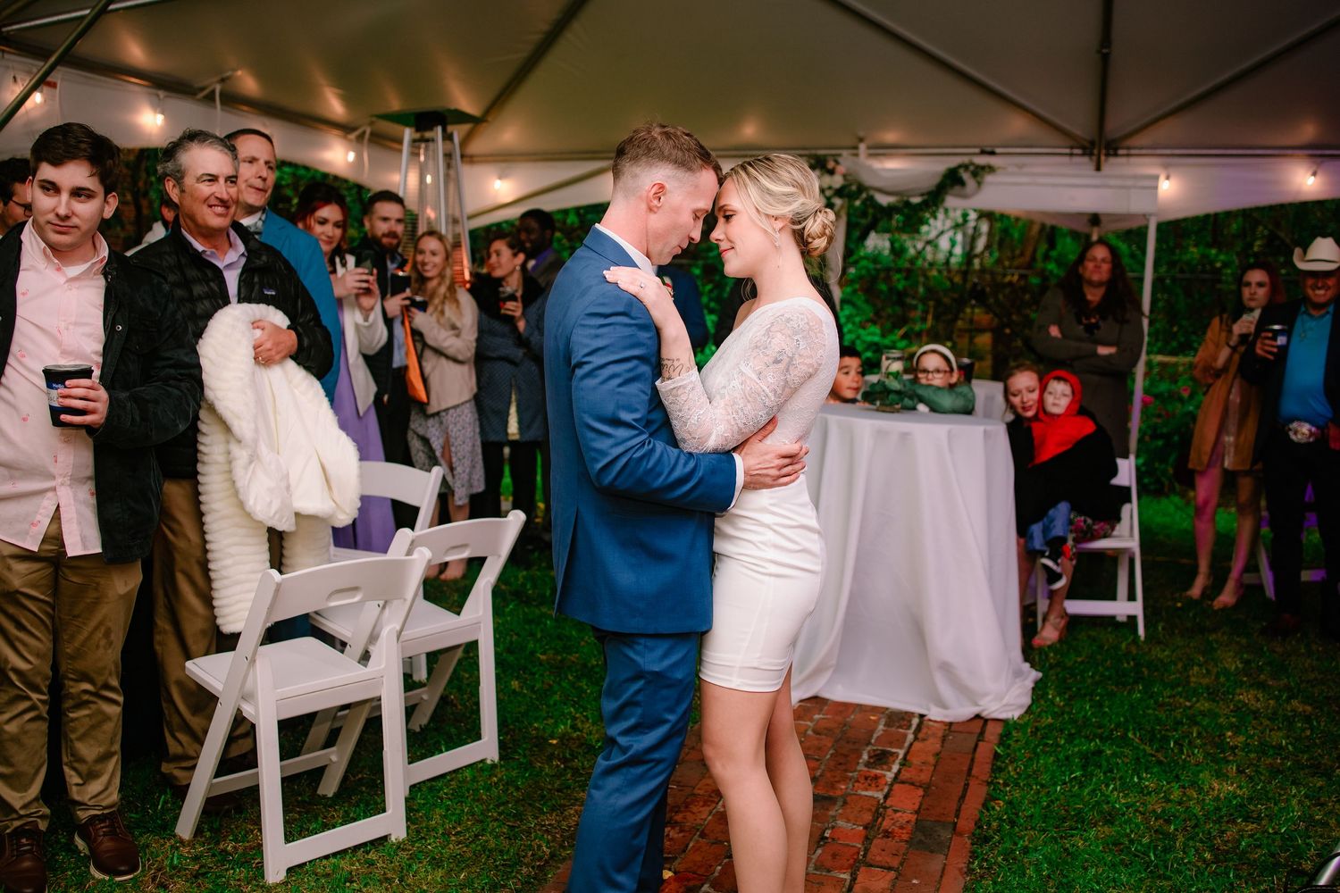 A couple shares their first dance under a tent at an outdoor wedding reception surrounded by smiling guests.