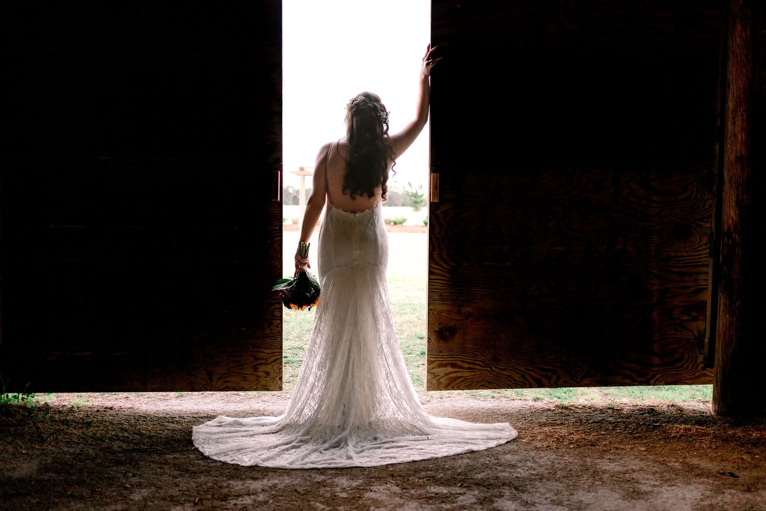 Bride in a lace wedding gown standing in barn doorway holding a bouquet in silhouette.
