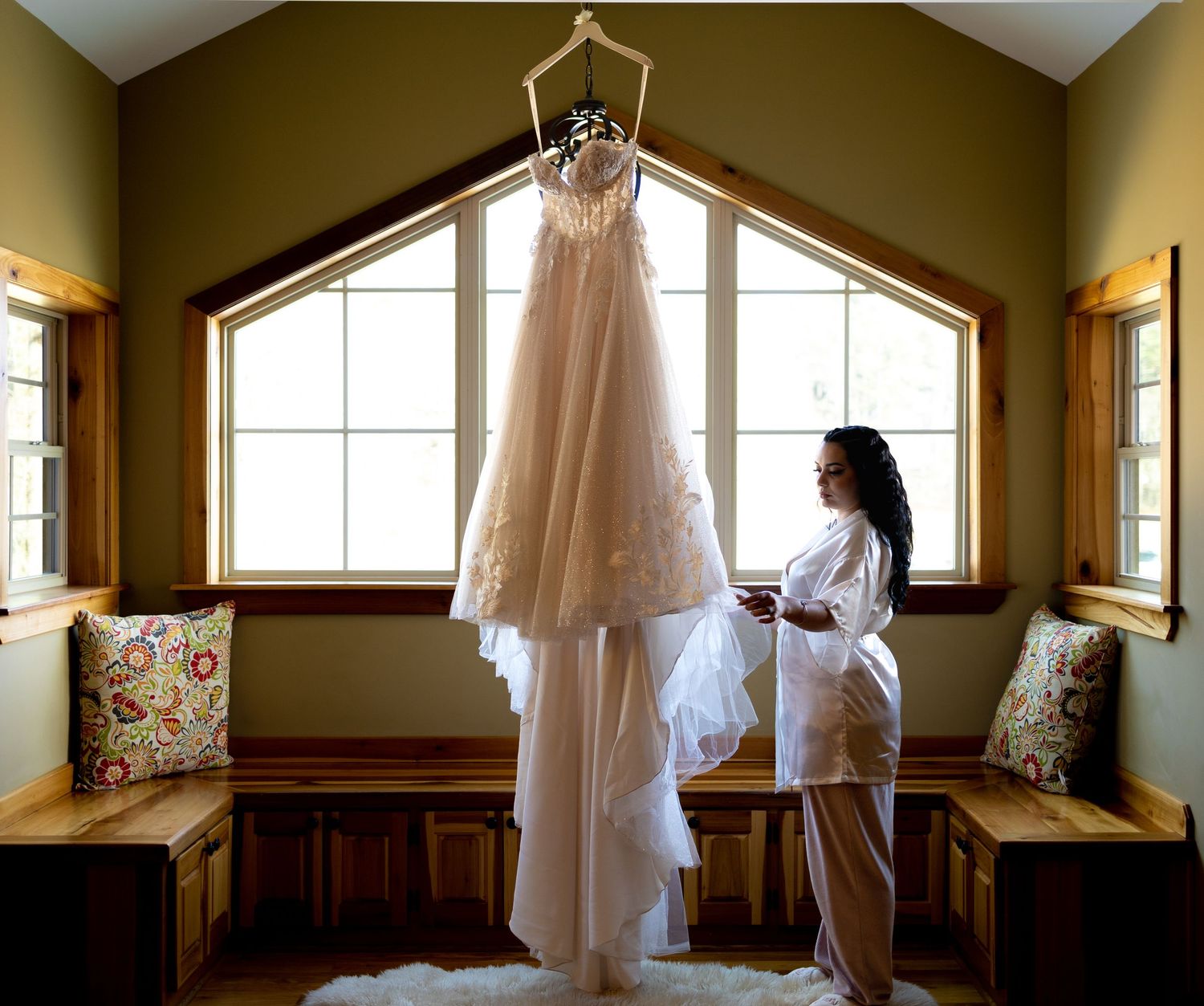 Bride in white robe admiring her elegant lace wedding dress hanging by a window in a cozy bridal suite.