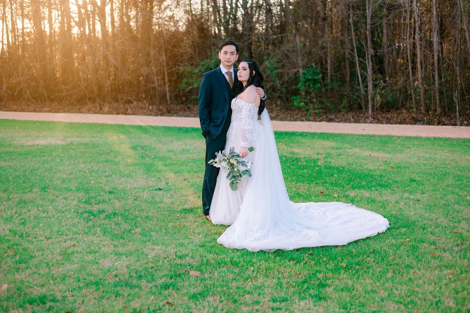 Bride and groom standing on lush green lawn at golden hour, bride in lace gown with long cathedral train.
