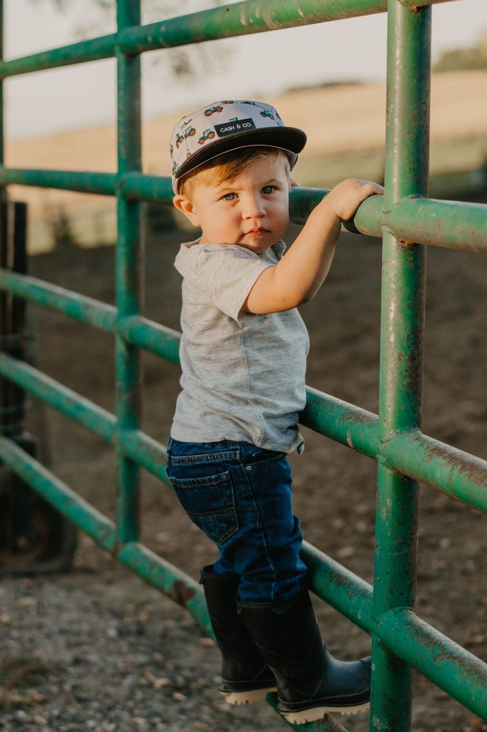 Toddler in baseball cap and boots climbing on green metal farm gates.