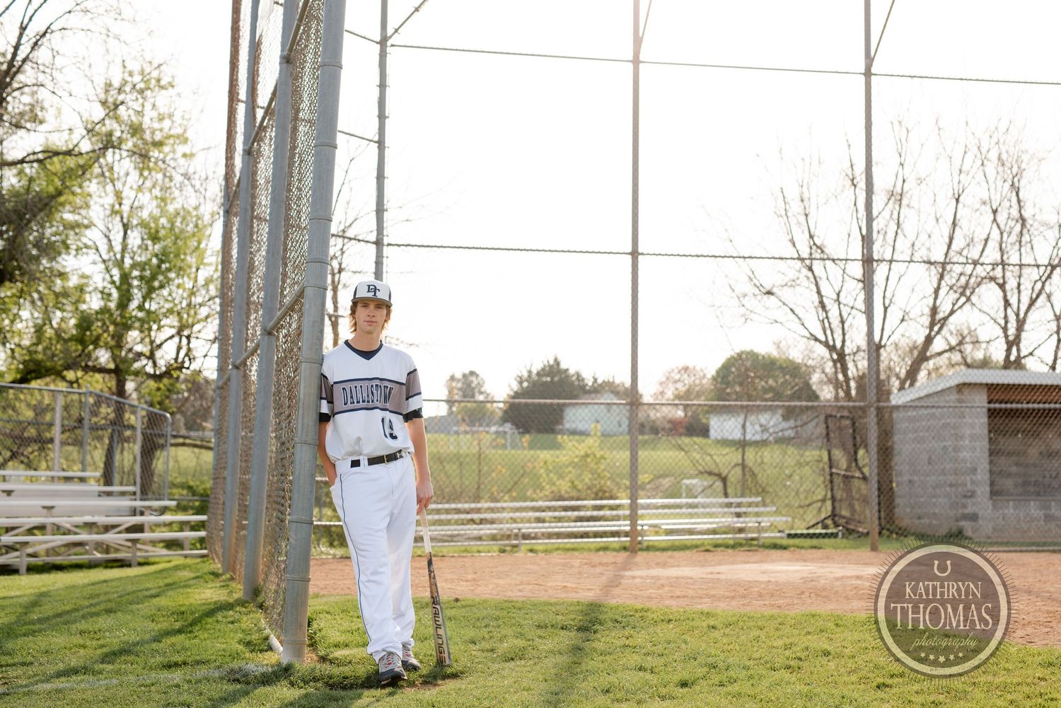 Baseball player stands beside fence pole on sunny field wearing white uniform.