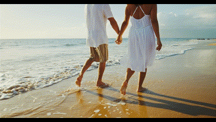 Two people walking hand in hand on a sandy beach at sunset with ocean waves visible.