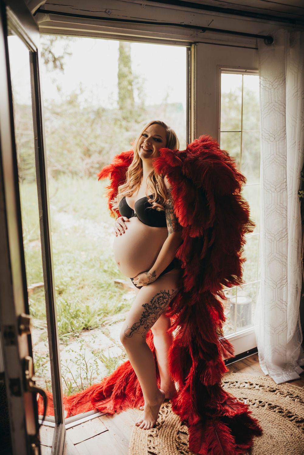 A woman poses in black lingerie and dramatic red feather boa near a bright window.
