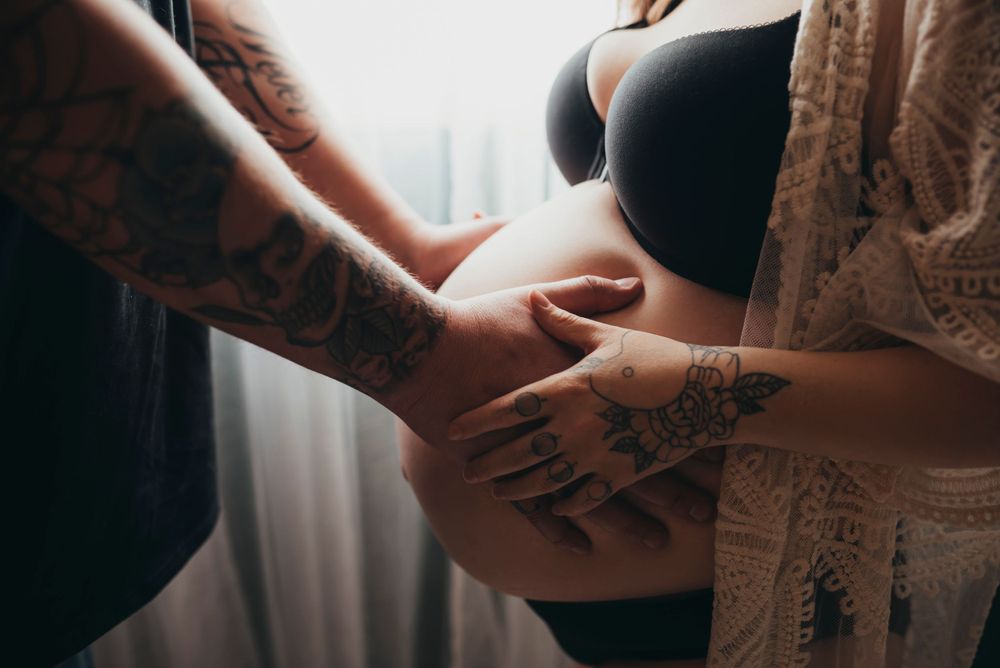 Close-up photo of hands with tattoos touching a pregnant belly in soft window light.