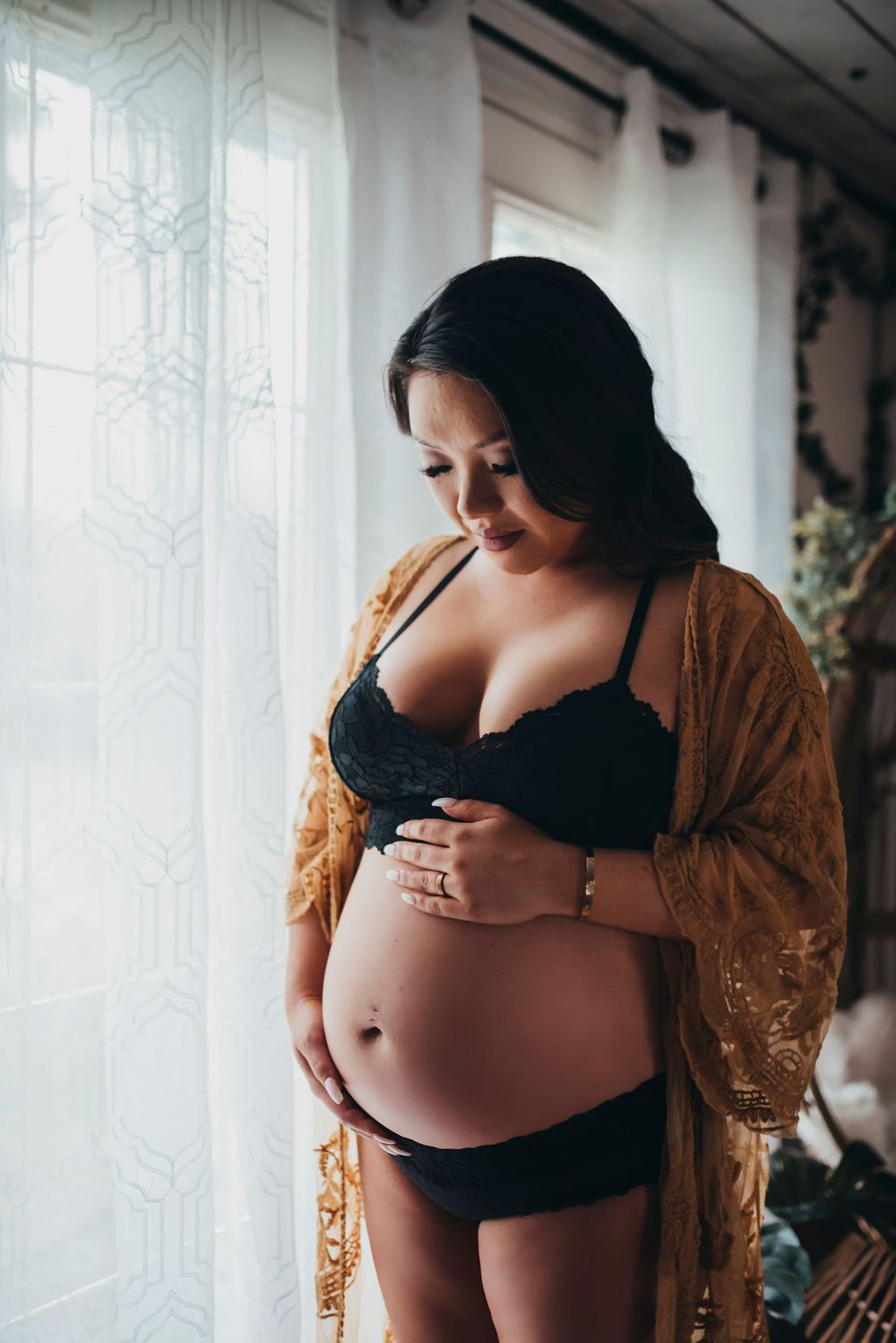 Expectant mother in black lingerie and gold lace robe poses by window in intimate maternity photoshoot.