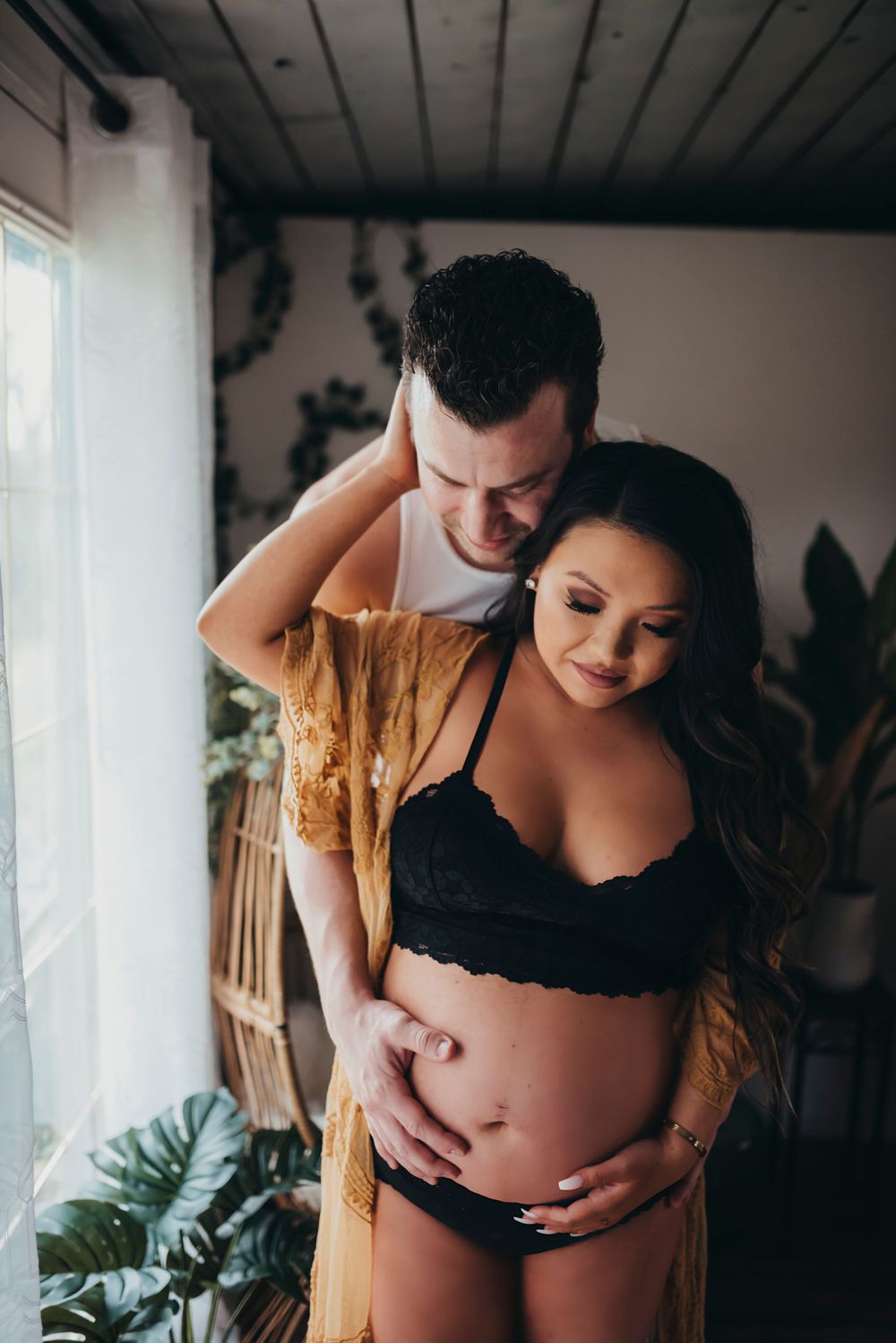 Couple embraces in intimate maternity photoshoot with warm lighting near window and indoor plants.