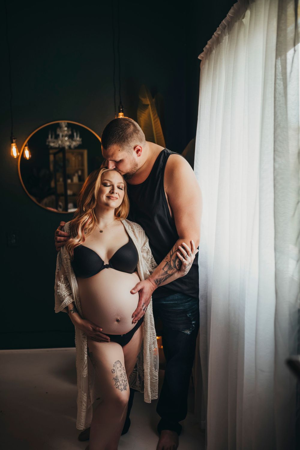 Intimate maternity photo featuring couple in dimly lit room with sheer curtains and romantic ambiance.