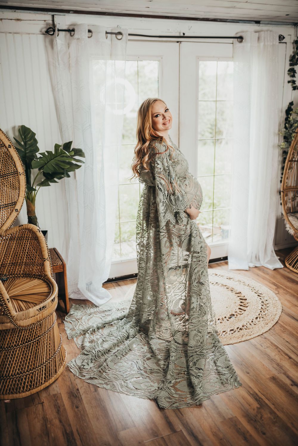 Elegant maternity photo in a flowing sage green lace dress standing by bright windows and wicker peacock chairs.