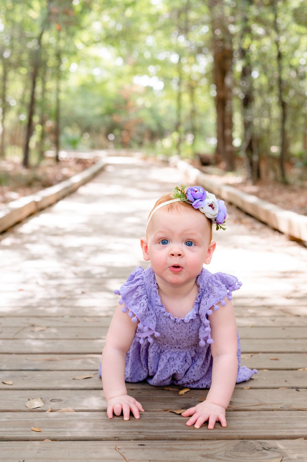 Baby in purple dress and floral crown crawls on wooden boardwalk through forest.