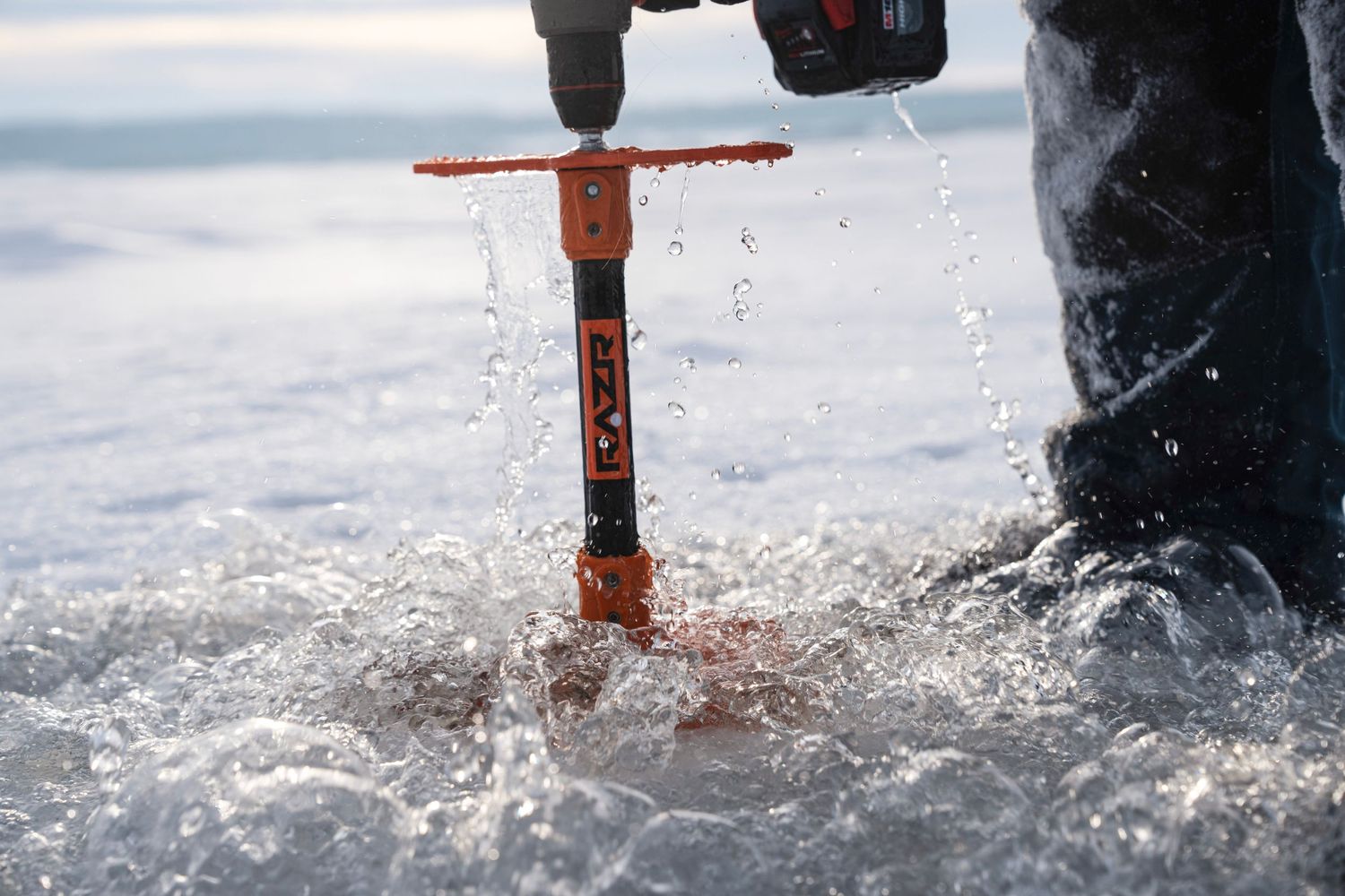 Close-up of an orange ice auger drilling through ice with snow spray.