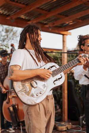 Musician playing a white resonator guitar under a wooden pavilion roof.