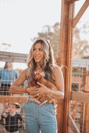 A woman in denim overalls holds a brown chicken in a rustic farm setting.