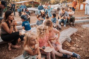 Children sit together on wood chips at an outdoor event enjoying entertainment.