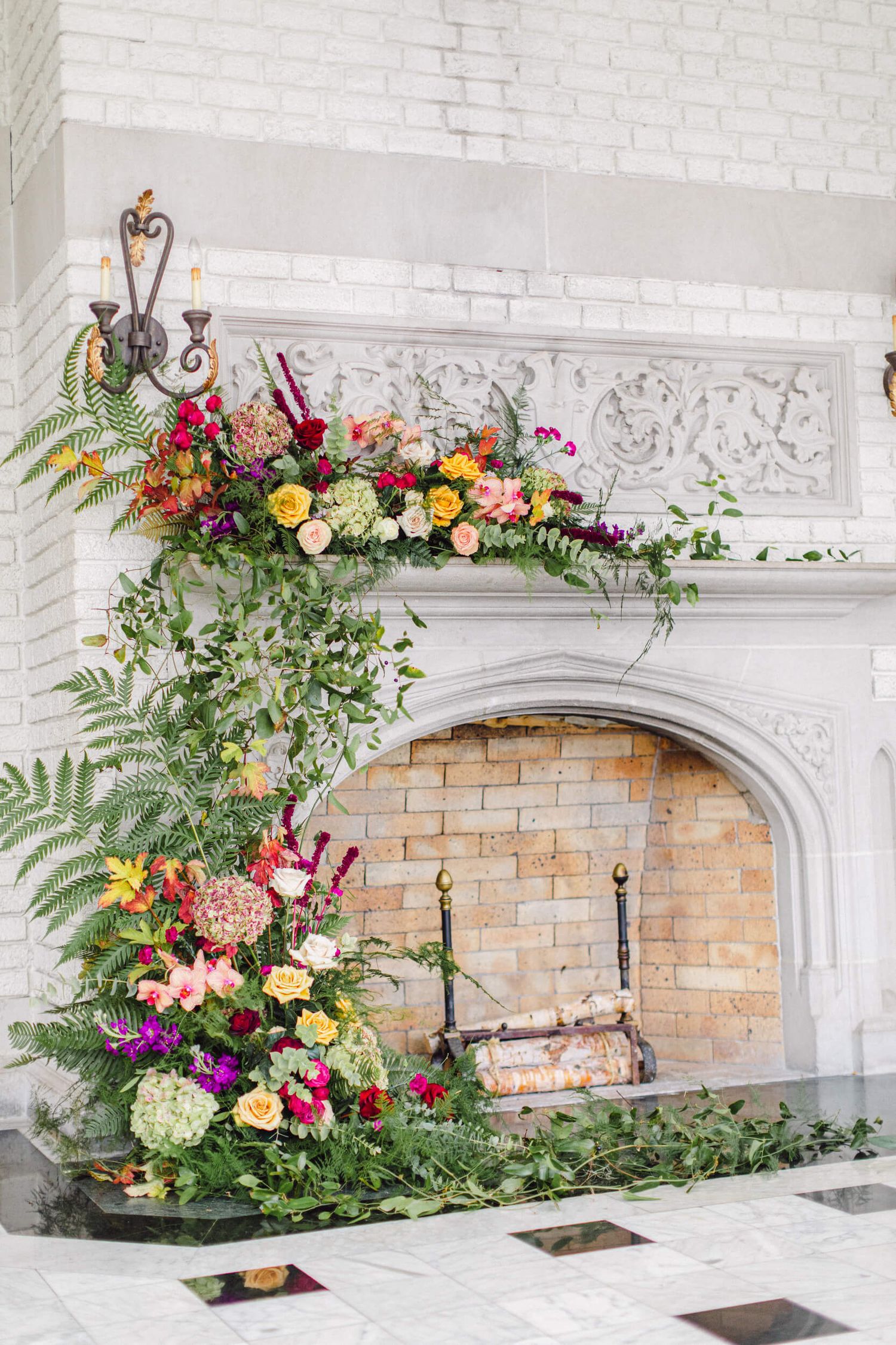 A lush floral arrangement with ferns and colorful blooms decorates a white brick fireplace.