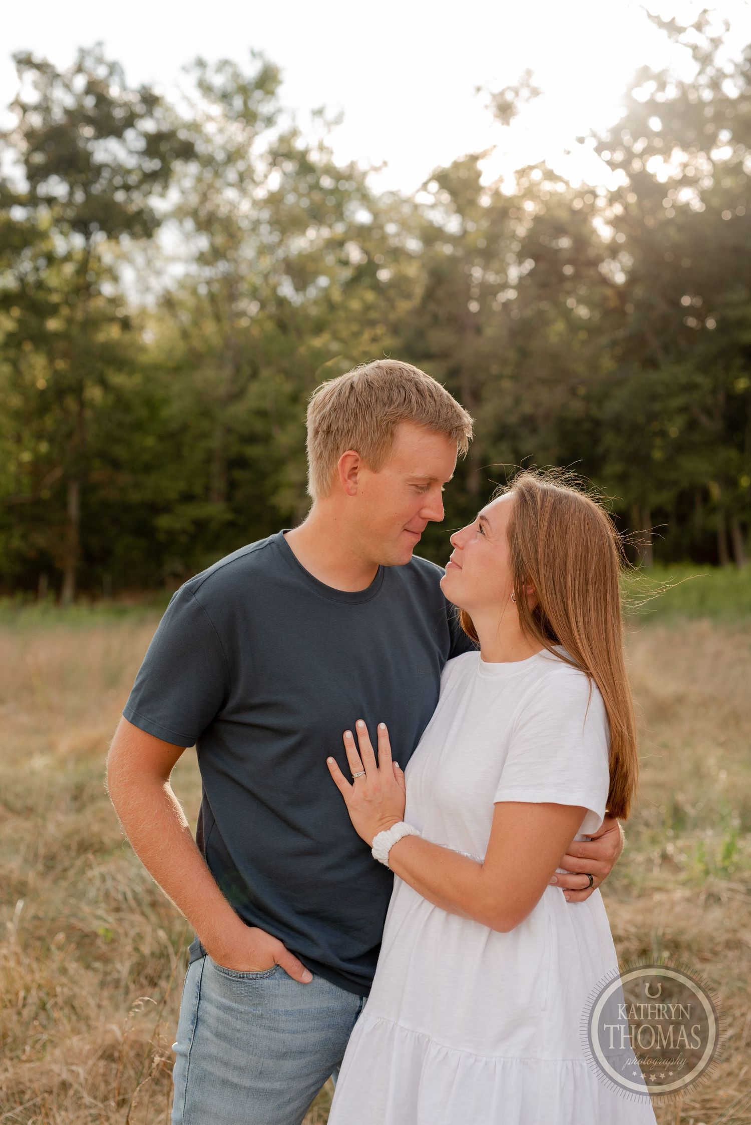 An intimate moment between a couple standing in tall grass with forest backdrop at sunset.