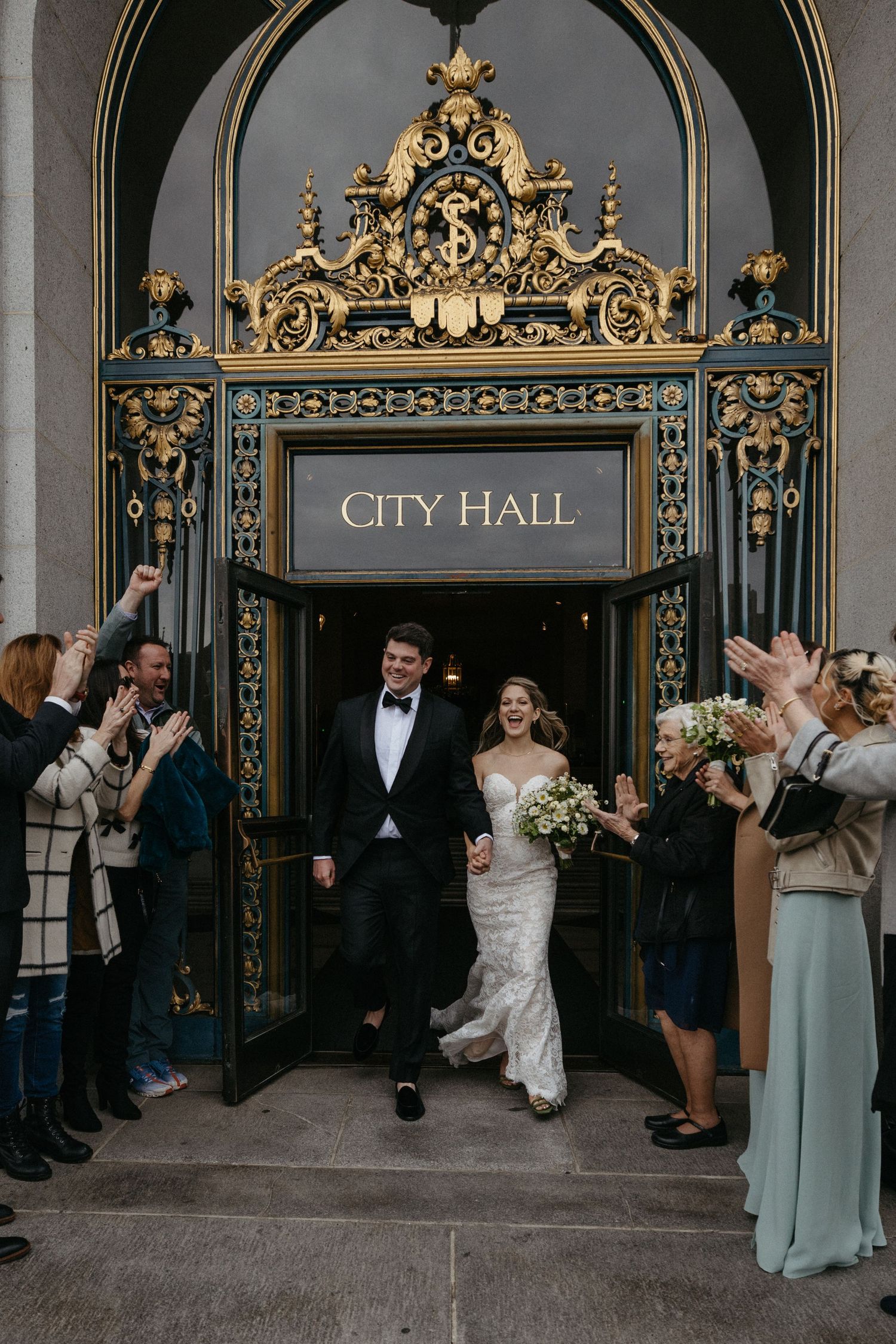 Newlyweds exit ornate City Hall doors while guests celebrate in formal attire.