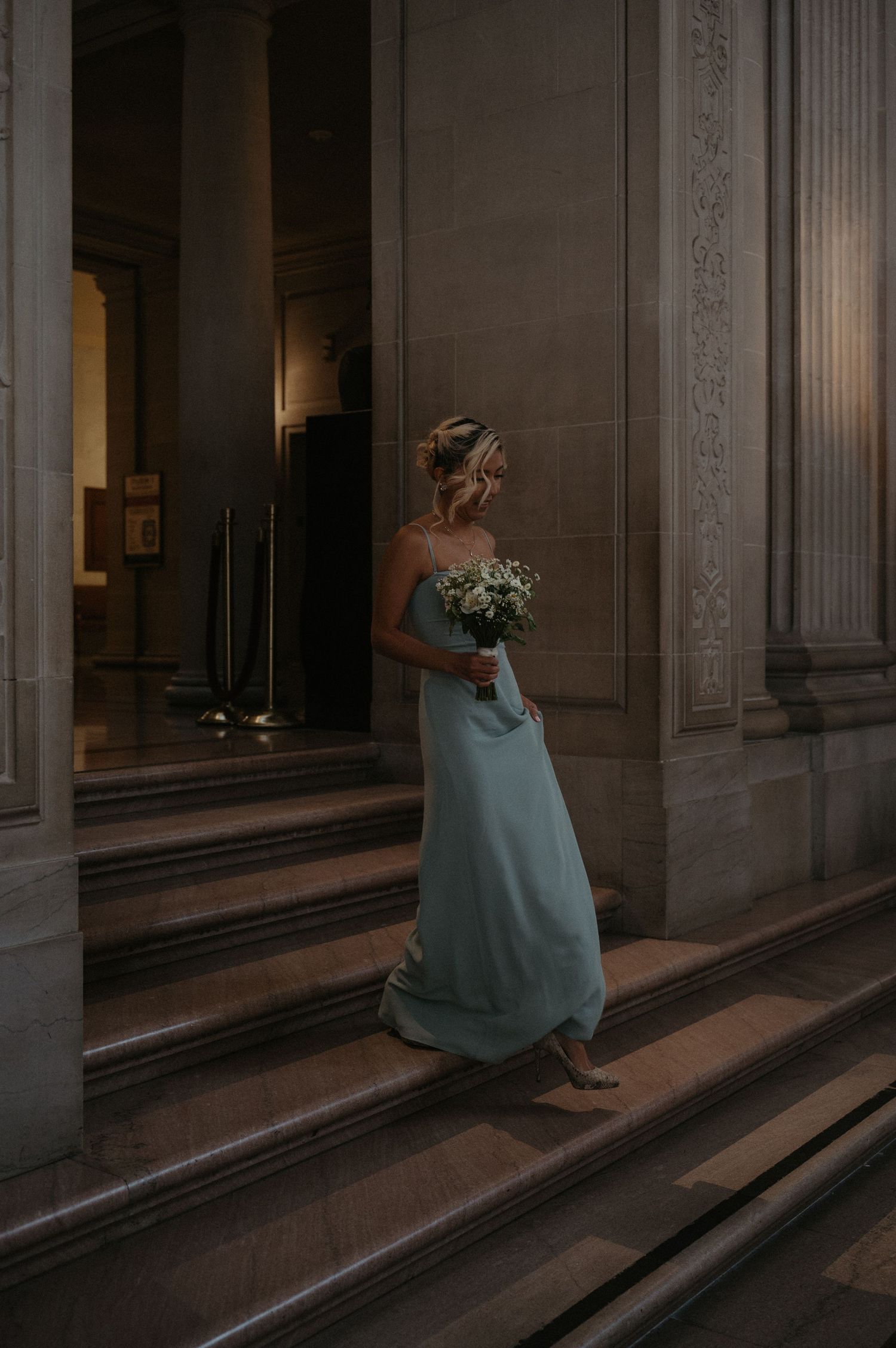A bridesmaid in a light blue dress carries a bouquet while ascending ornate stone steps.