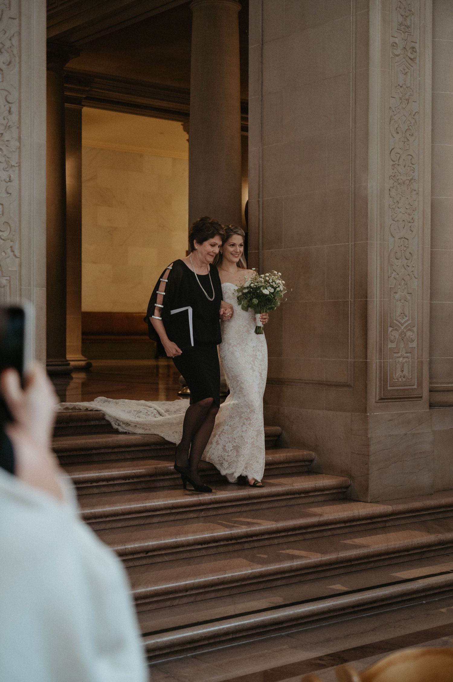 A couple descends grand stone steps in formal wedding attire while guests capture photos.