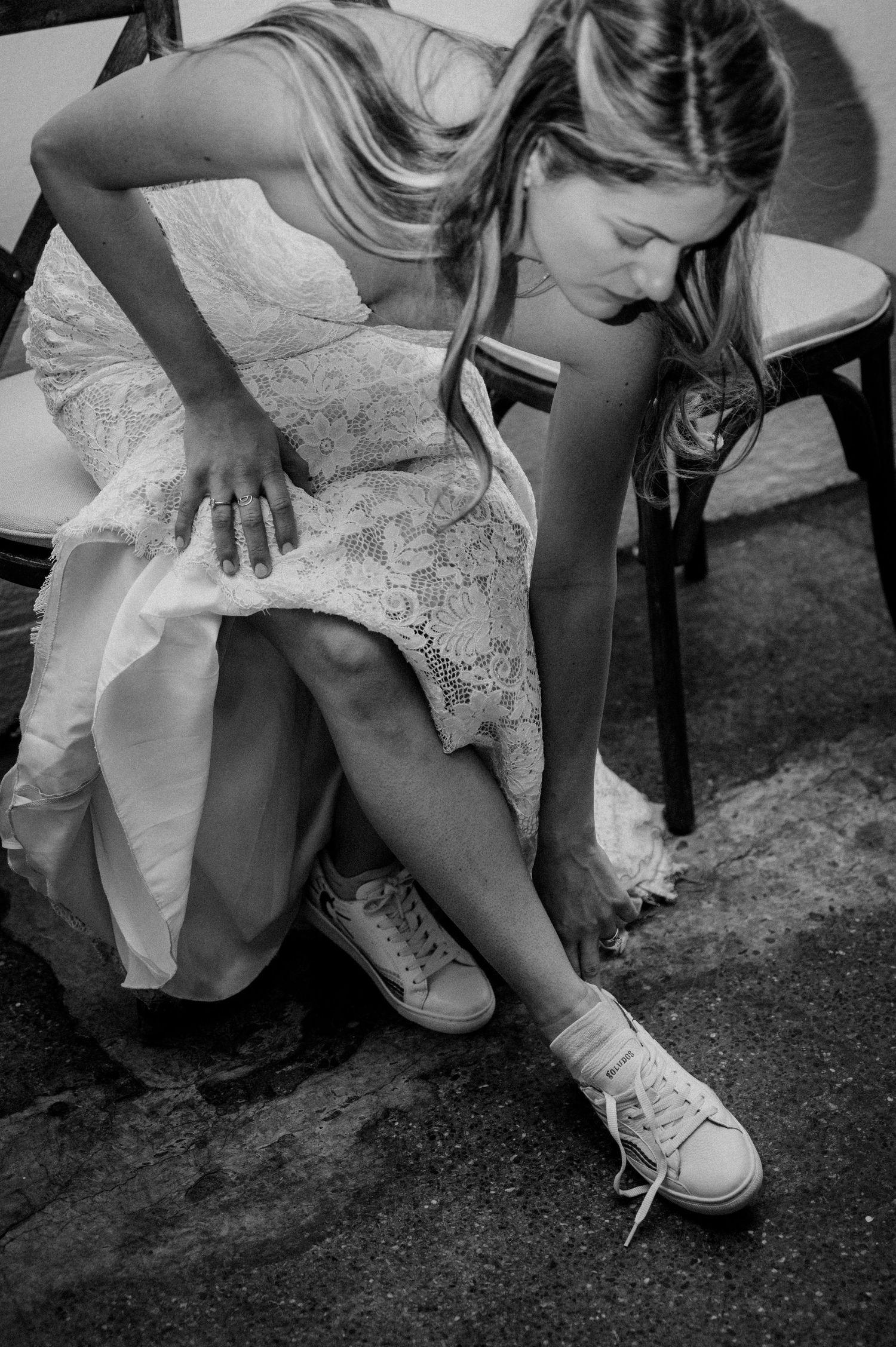 A bride in a lace dress adjusts her casual sneakers before the reception.
