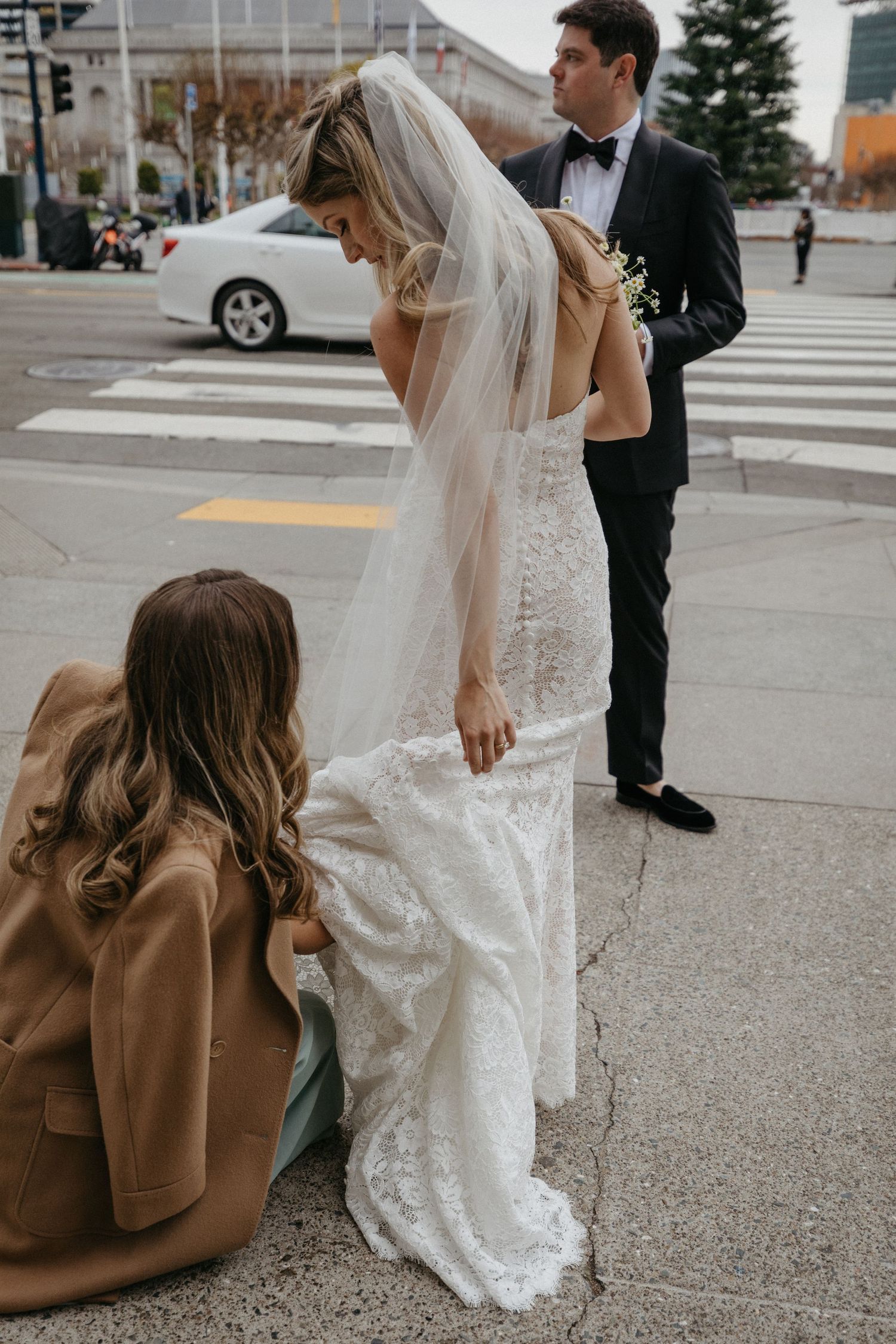 Helper adjusts wedding dress train on city sidewalk as couple prepares for photos.