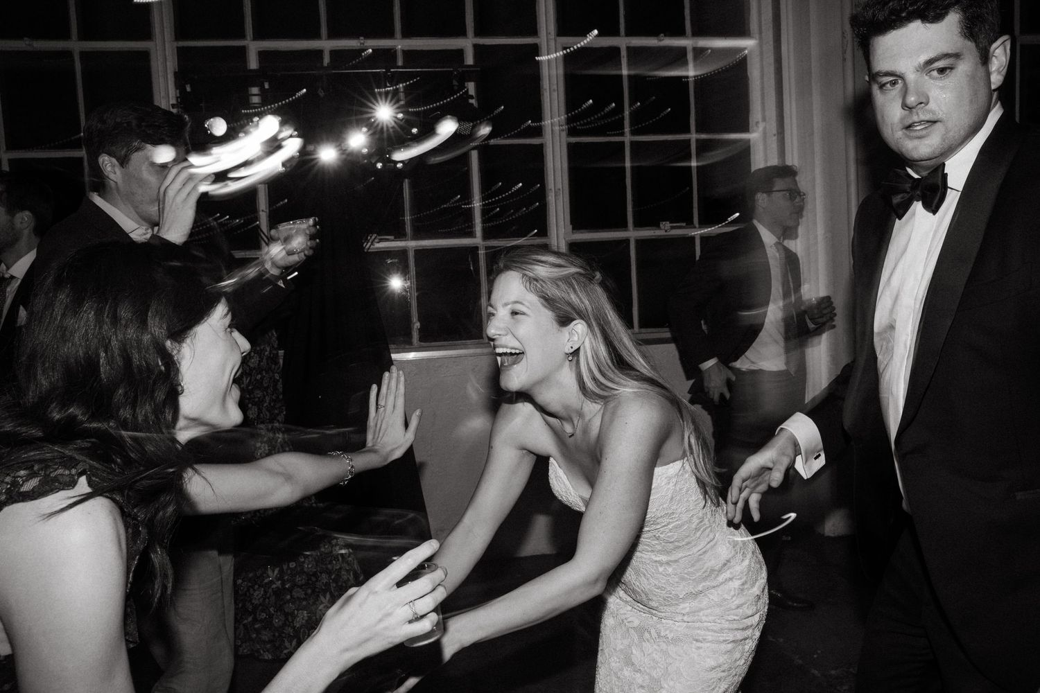 Wedding guests dance and celebrate on a dance floor near large windows at night.