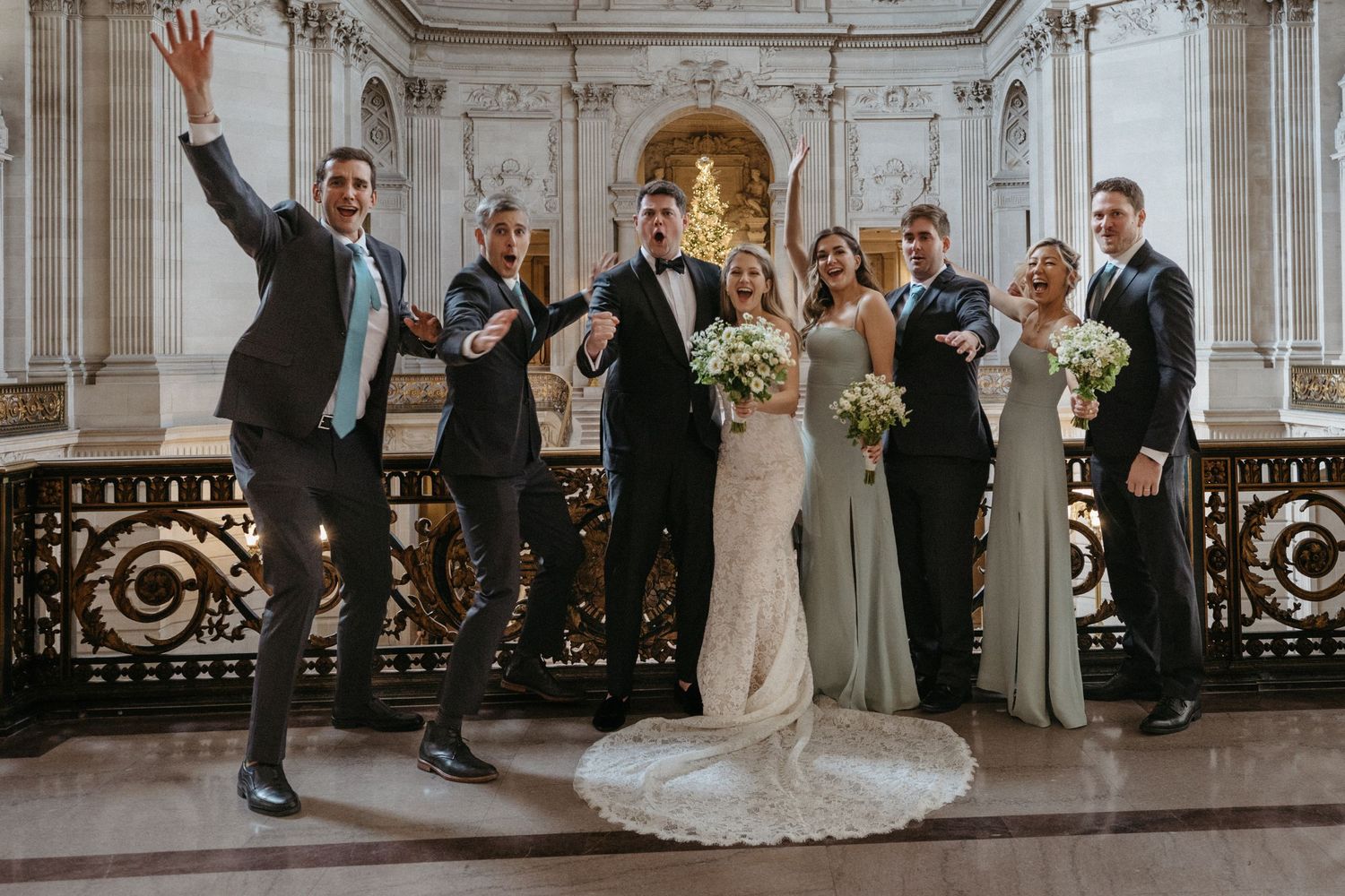 Wedding party celebrates joyfully on marble staircase in front of illuminated golden archway.