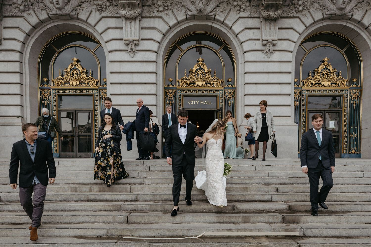 Wedding party descends ornate City Hall steps with decorative gold arches in background.