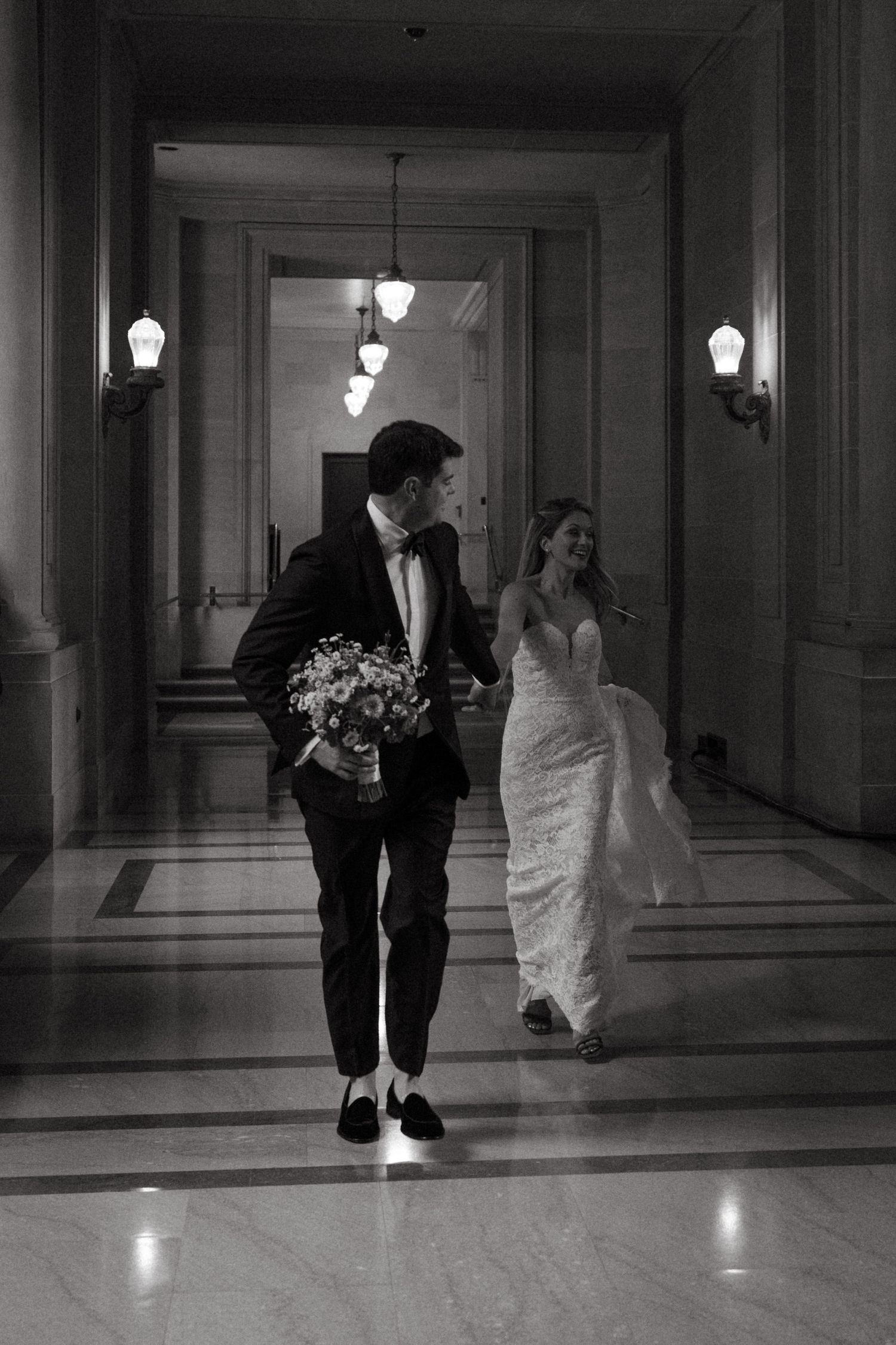 A couple walks through an elegant hallway with classic wall sconces and marble floors.