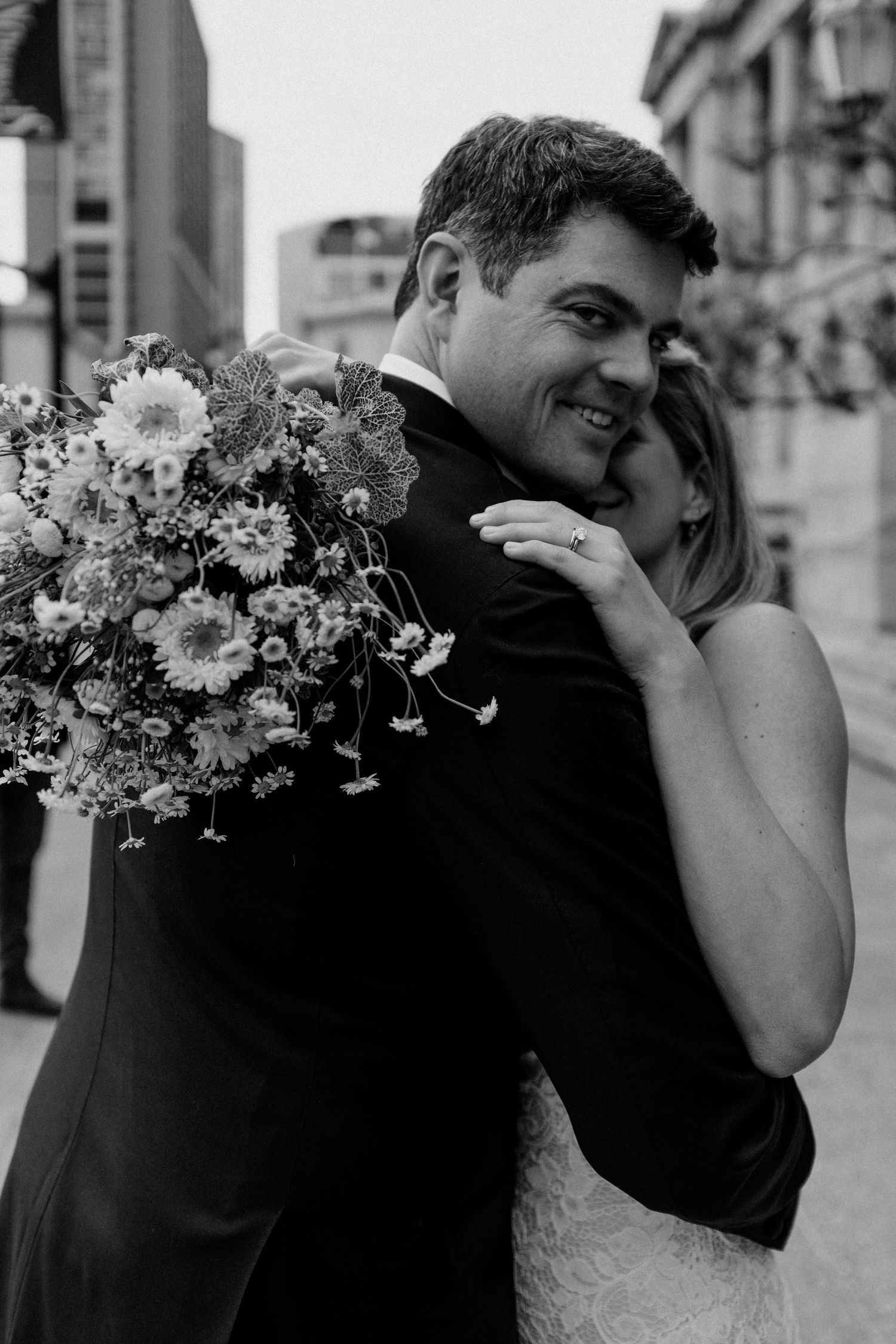 Black and white portrait of newlyweds embracing with floral bouquet in urban setting.