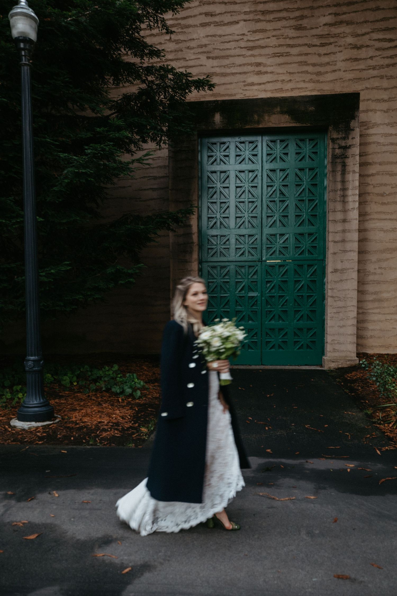 A person in a wedding dress and long coat holds a bouquet while standing in front of a teal decorative door.