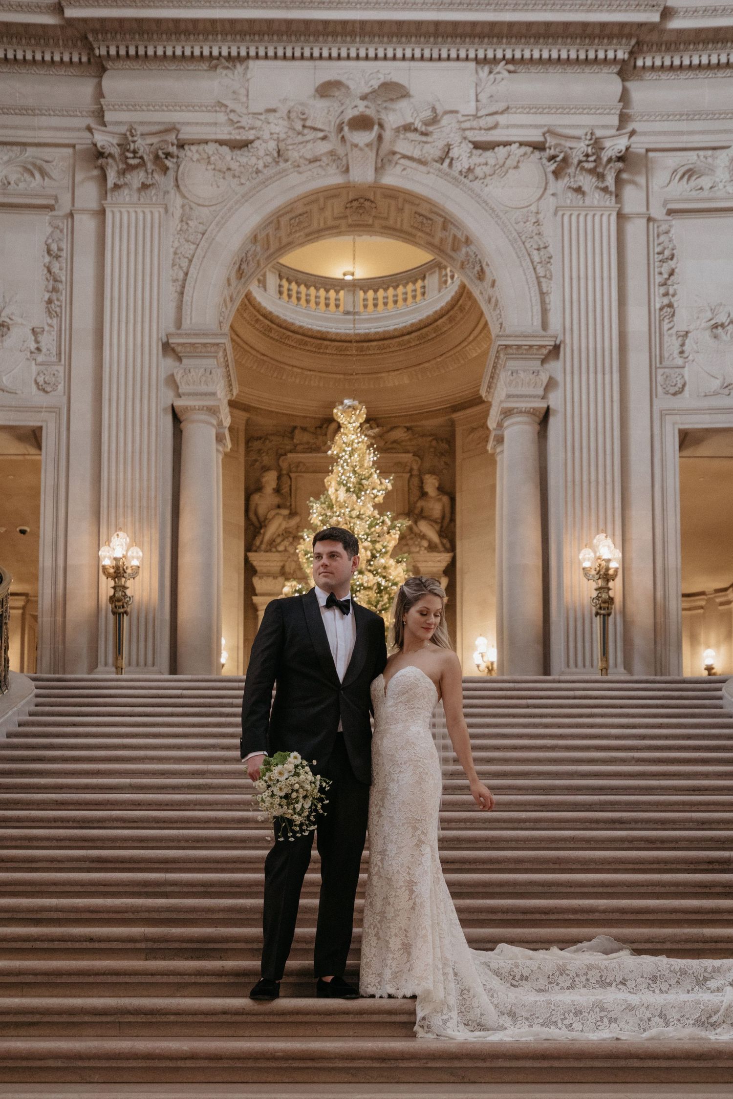 A wedding portrait on marble steps with a glowing Christmas tree and ornate architecture in the background.