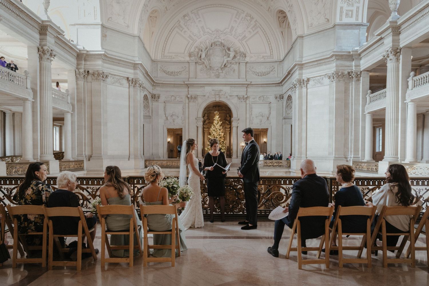 Wedding ceremony in a grand white hall with vaulted ceilings and wooden chairs.