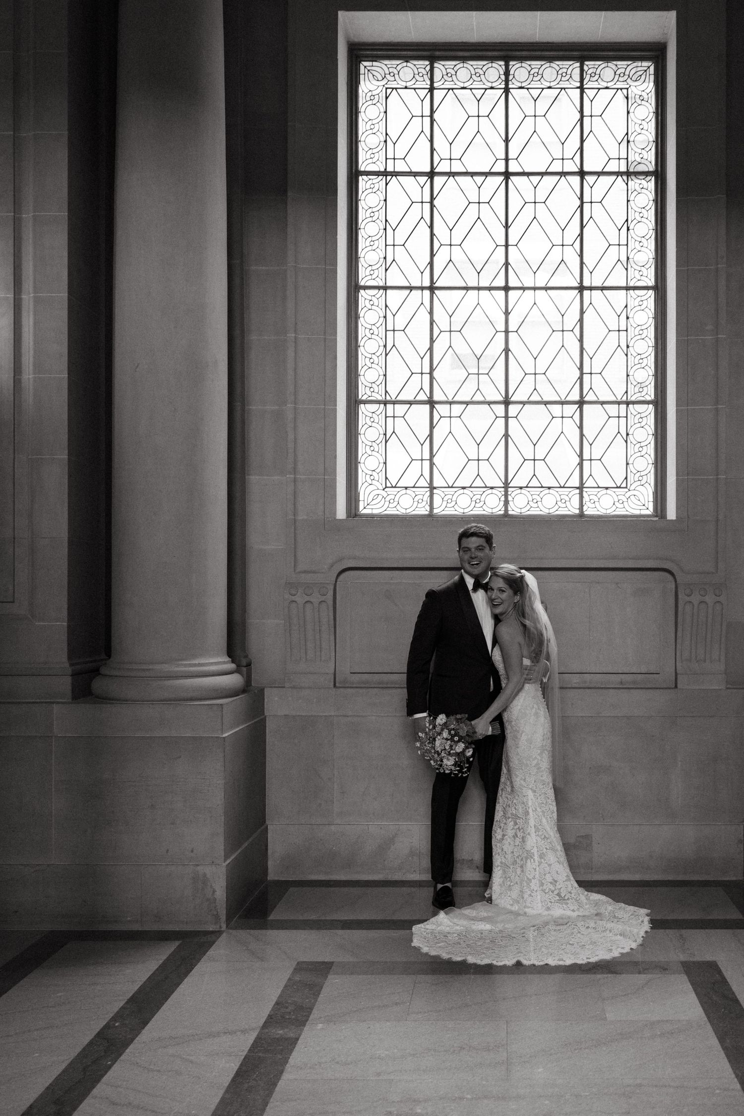 A wedding couple poses elegantly in front of a grand window with geometric patterns in a formal interior.