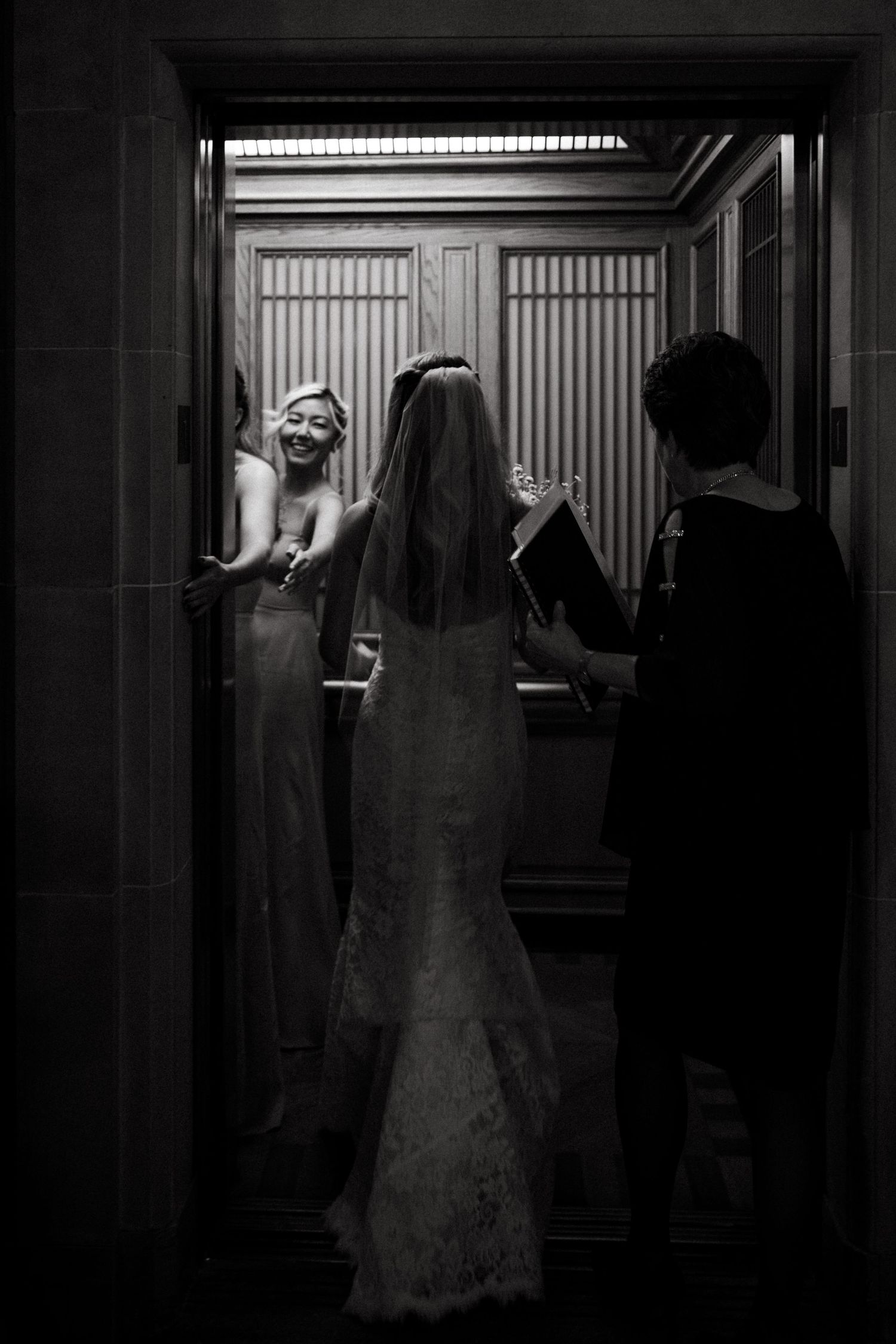 Black and white photo of a bride in a wedding gown looking in an elevator mirror.