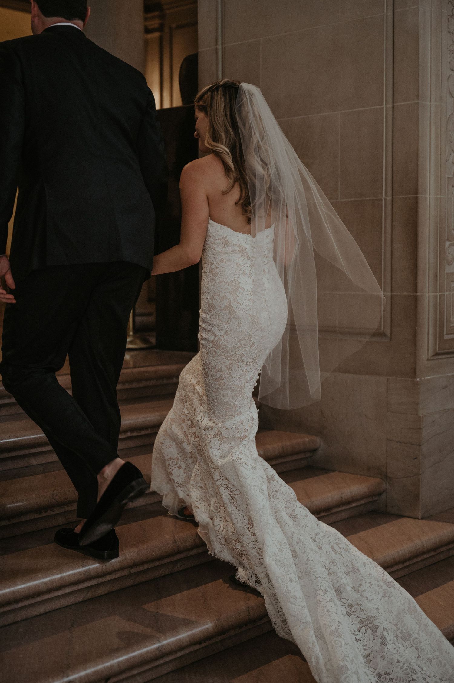 A couple descends stairs during their wedding, showing off the bride's flowing lace gown and long train.