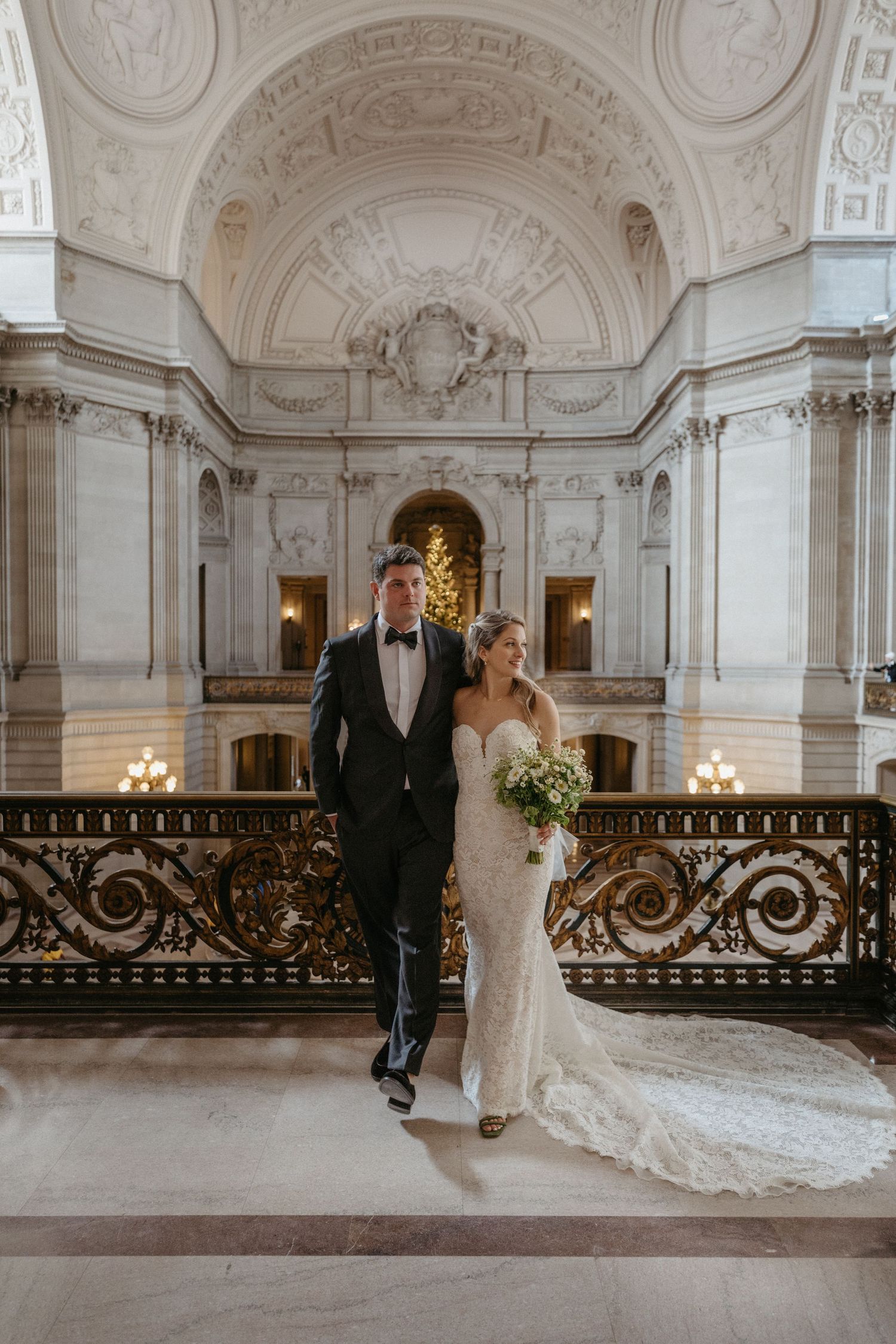 A wedding couple stands in front of a beautifully lit archway in an ornate marble hall.
