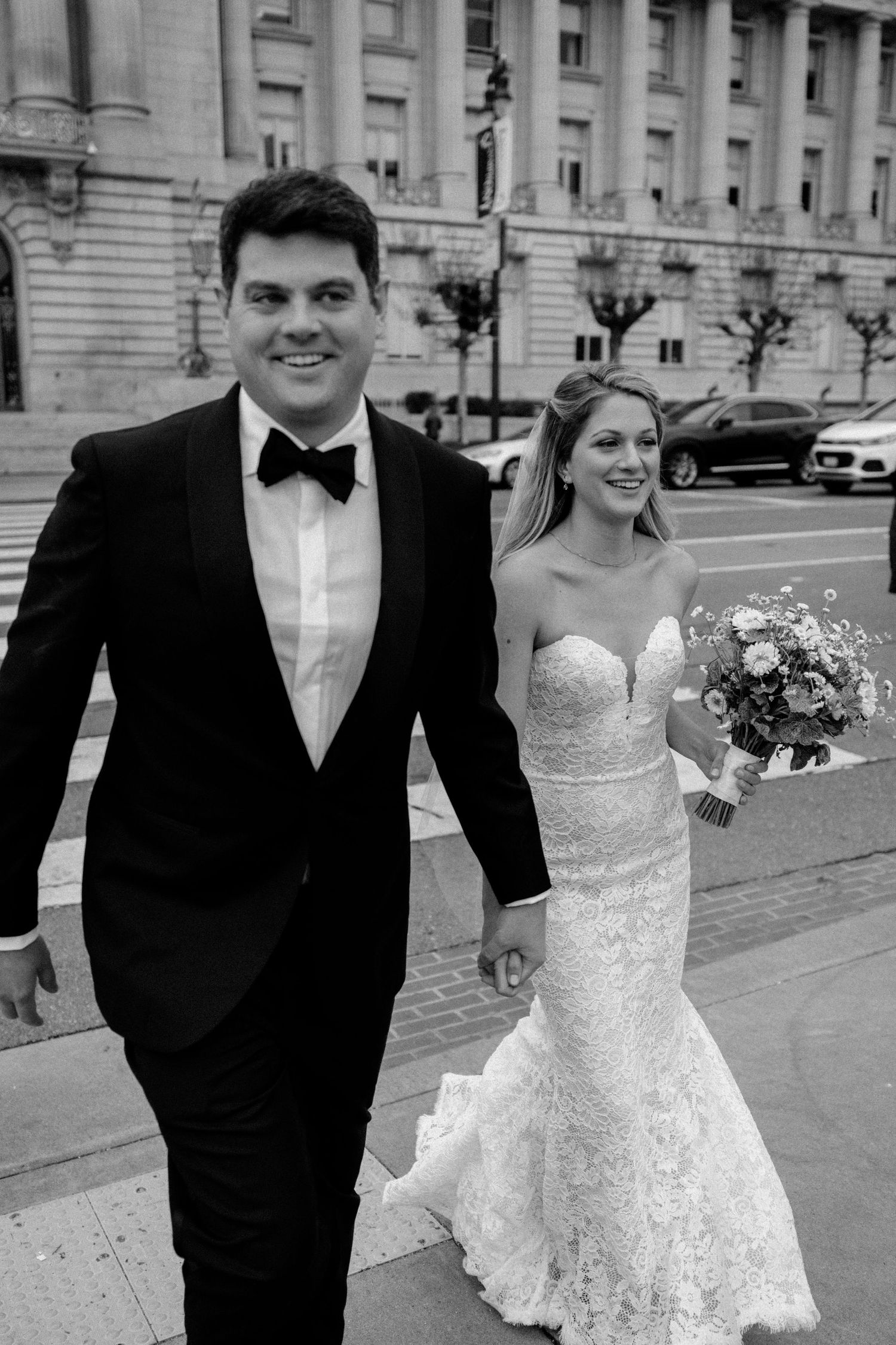 Black and white portrait of newlyweds walking along city sidewalk after ceremony.
